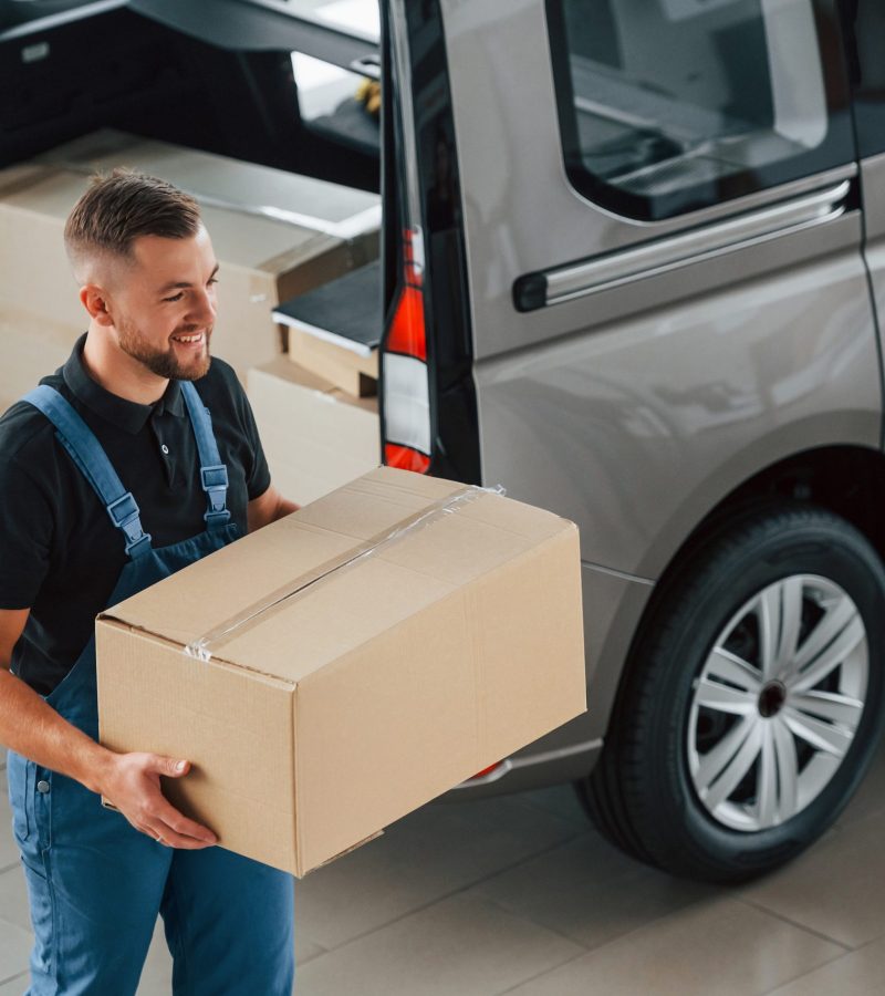 Walking and holding box in hands. Delivery man in uniform is indoors with car and with order.